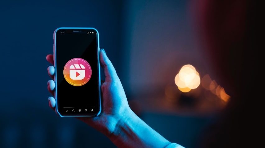 A imagem mostra uma mão segurando um smartphone que exibe o ícone de um aplicativo de vídeo, com um design que combina cores rosa e laranja. O fundo é suavemente iluminado em tons de azul, criando uma atmosfera moderna e envolvente. A tela do telefone é escura, destacando o ícone centralizado, que parece ser uma representação de um editor de vídeo ou uma plataforma de compartilhamento de vídeos. A mão que segura o dispositivo é visível, com um toque delicado, sugerindo que a pessoa está prestes a interagir com o aplicativo. A iluminação suave e o foco no smartphone transmitem uma sensação de tecnologia e criatividade.  A  imagem representa exemplos de como fazer reels