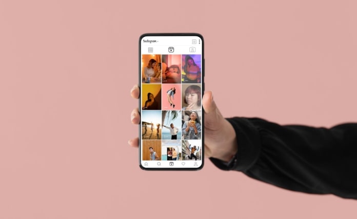 A imagem mostra uma mão segurando um smartphone que exibe a interface do Instagram. A tela do dispositivo apresenta uma grade de fotos, com várias imagens de pessoas em diferentes poses e ambientes, sugerindo um conteúdo diversificado e visualmente atraente. O fundo da imagem é de uma cor rosa suave, que contrasta com a tela do telefone. A mão que segura o smartphone parece estar vestida com uma manga preta, indicando um estilo casual. A composição transmite uma sensação moderna e conectada, típica das interações nas redes sociais.  A  imagem representa exemplos de como fazer reels
